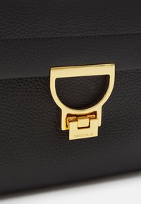 Sort lædertaske med en tekstureret overflade, udstyret med et guldmetallåg og lås. Logoet "Coccinelle" er graveret på låsen.