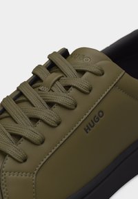 Baskets en cuir vert olive avec lacets assortis, semelle noire, et logo "HUGO" embossé sur le côté et la languette.