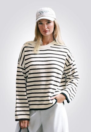 Pull rayé crème et noir, avec une coupe décontractée, des poignets et un ourlet côtelés. Assorti à un pantalon clair et une casquette blanche.