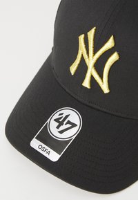 Melnā beisbola cepure ar izšūtu dzeltenu "NY" logotipu. Tam ir izliekts sēžamā daļa un izmēra uzlīme ar norādi "47" un "OSFA." Tekstūrēta auduma.