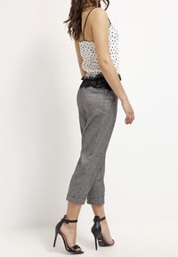 Top in cotone bianco a pois neri con bordatura in pizzo nero, abbinato a pantaloni cropped grigi. Il modello indossa décolleté con cinturino alla caviglia neri, evidenziando un design pulito.