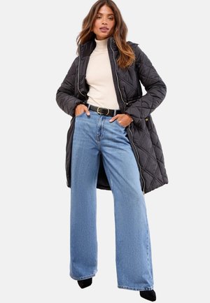 Gequilted zwarte lange jas met een capuchon, beige turtleneck en wijde blauwe jeans. Zwarte riem met een gouden gesp. Zachte texturen en opvallende vormen.
