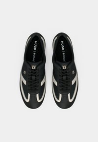 Ténis pretos com parte superior texturizada, detalhes em creme e costuras brancas contrastantes. Incluem fecho de atacadores e marca no língua.