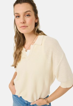 Pullover a maglia color beige chiaro con scollo a V e maniche corte. Tessuto strutturato con una vestibilità comoda. Indossato sopra una maglietta bianca, abbinato a jeans blu.