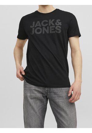 Mann trägt schwarzes "JACK & JONES" T-Shirt und graue Jeans, zieht das T-Shirt leicht mit der linken Hand, vor hellem Hintergrund isoliert.