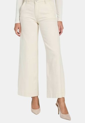 Donna che indossa pantaloni crema a vita alta e gamba larga abbinati a décolleté beige con punta appuntita, in piedi su sfondo bianco.