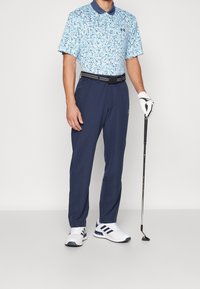 Pánský golfový outfit obsahující světle modrou polo košili s květinovým vzorem, námořnické kalhoty, černý opasek a bílé boty s tmavými akcenty, držící golfovou hůl.