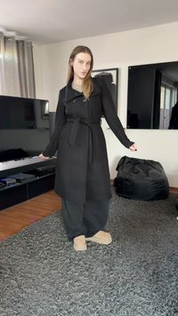 Pikk must mantel vööga, topeltrinnaline disain ja nööbiaktsendid. Kombineeritud hallide pükste ja beežide sussidega, tekstuurse vaiba peal.