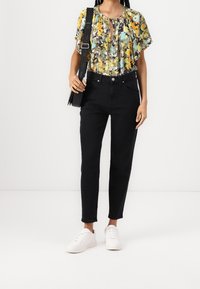 Žena v černých vysokých culottes jeans, bílých teniskách, květované blůze s krátkým rukávem a s černou crossbody taškou s značkovým popruhem.