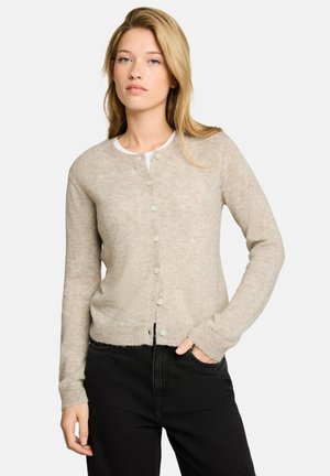 Junge Frau mit blonden Haaren, die eine beige geknöpfte Strickjacke und schwarze Hose trägt, steht mit einer Hand in der Tasche vor schlichtem Hintergrund.