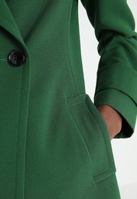 Main d'une personne dans la poche d'un manteau vert sur mesure avec un grand bouton noir et une manche retroussée sur fond clair.