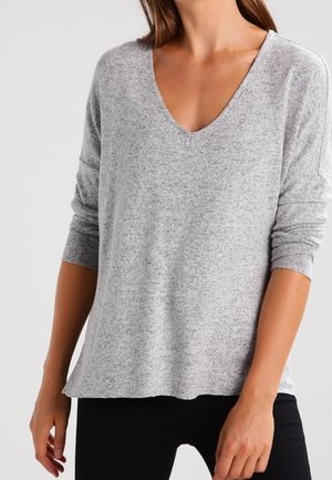Femme portant un pull gris clair à manches longues et col en V, avec un pantalon noir, debout avec les bras détendus le long du corps.