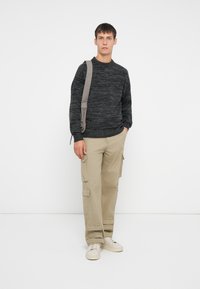 Ung man som bär en mörkgrå texturerad tröja, beige cargobyxor, vita sneakers och en grå ryggsäcksrem över ena axeln.
