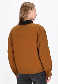 Brun, let jakke med en kordfløjlskrage, elastisk kant og knapmanchetter. Glat tekstur, afslappet design og oversized pasform.
