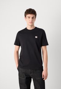 Zwarte t-shirt met korte mouwen van een soepel materiaal, voorzien van een klein rood en wit logo op de borst. Gekleed met donkere spijkerbroek.