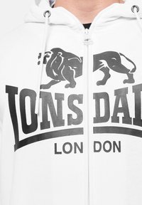 White zip-up hoodie featuring a black lion graphic and bold text reading "LONSDALE LONDON." Made of soft fabric with drawstring hood.