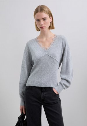 Suéter gris con cuello en V adornado con encaje. Mangas largas, textura acanalada y cuerpo ajustado combinado con jeans negros.