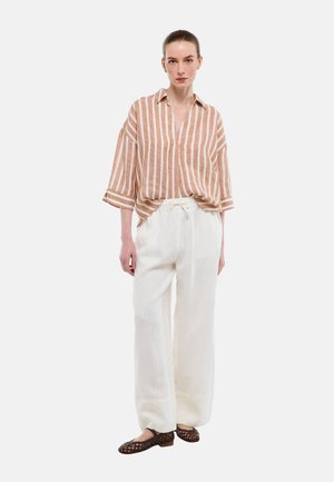 Femme portant une chemise ample à rayures marron et blanches, un pantalon large blanc et des chaussures plates tressées foncées, debout devant un fond blanc.