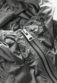 Könnyű szürke nylon dzseki elülső cipzárral, elasztikus részletekkel és logós címkékkel. Texturált felület finom fényléssel.