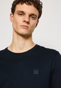 Camiseta de manga larga de color azul marino hecha de algodón, con un pequeño parche de logo cuadrado negro en el lado izquierdo del pecho, cuello redondo y corte estándar.