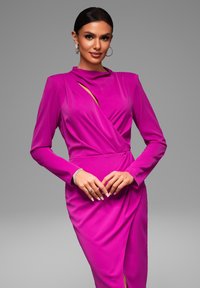 Getailleerde jurk met lange mouwen van levendig roze stof. Beschikt over een gedrapeerd ontwerp met een asymmetrische halslijn en een zijsplit. Elegante oorbellen en armband.