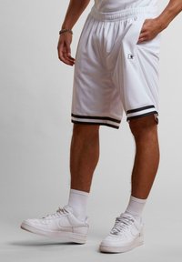 Shorts de sport blancs avec des rayures noires au bas, taille élastique et détail de logo sur la cuisse. Portés avec des baskets blanches et des chaussettes.