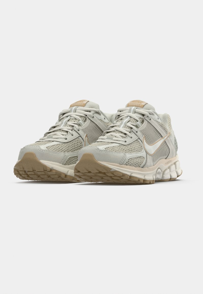 Hellgraue Sportschuhe mit Mesh-Obermaterial, Schnürverschluss, beigen Akzenten und strukturiertem Gummiauslauf. Auffälliges Nike-Logo in Weiß.