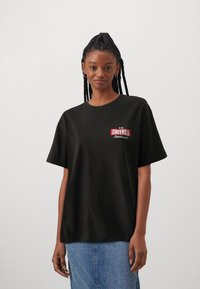 Sort bomulds t-shirt med korte ærmer, der har et rødt og hvidt "ZOYORK" logo på fronten, parret med blå denimbukser.