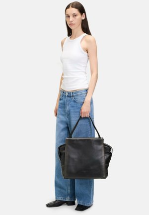 Schwarze Leder-Tasche mit doppelten Griffen, strukturiertem Design, glatter Textur und seitlichen Akzenten, gehalten von einer Person in legerer Kleidung.