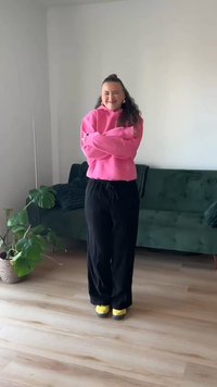 Hanorac roz, pantaloni largi negri și pantofi galbeni. Stând cu brațele încrucișate, zâmbind. Mediu interior cu canapea verde și plantă.