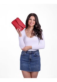 Červená kožená kabelka s klopou a texturovaným povrchem, držena v ruce ženy na jednobarevném bílém pozadí.