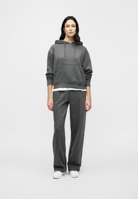 Γκρι φούτερ με κουκούλα, εμπρός τσέπη και κορδόνια, συνδυασμένο με γκρι παντελόνια φαρδιάς γραμμής που ταιριάζουν. Το μοντέλο φοράει λευκά αθλητικά παπούτσια.