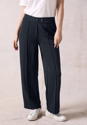 Pantalones amplios negros con una textura suave, pliegues frontales y una cintura elástica. Combinados con zapatillas blancas.