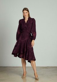 Rochie tip wrap, de un burgundy profund, cu modele florale texturate, cu mâneci lungi strânse și o tiv flared. Asortată cu toc înalt împodobit.