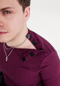 Burgundi színű ing, fekete tépőzárral a vállnál, sima textúrával. Ezüst lánc nyaklánc látható a nyak körül.