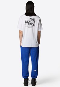 Camiseta blanca de algodón con texto gráfico negro en la parte posterior, combinada con pantalones de chándal azul con puños elásticos y logo, usados con calzado negro.
