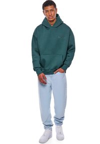 Grüne Kapuzenjacke mit großer Kapuze, Kängurutasche und gesticktem Logo. Kombiniert mit hellblauen Jeans und weißen Sneakern.