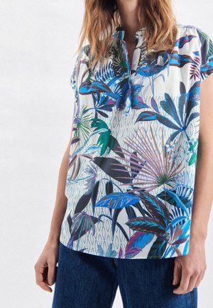 Kobieta ubrana w białą bluzkę z krótkim rękawem z kolorowymi niebieskimi, zielonymi i fioletowymi tropikalnymi wzorami liści oraz niebieskie dżinsy.