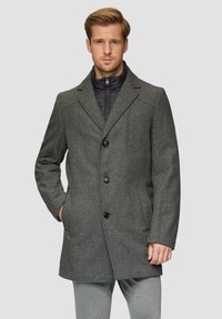 Grau karierten Übermantel mit abgerundetem Kragen, drei schwarzen Knöpfen und Seitentaschen, über einer schwarzen gesteppte Innenjacke getragen.