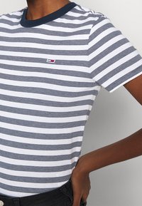 Person som bär en marinblå och vit horisontellt randig t-shirt med en liten rektangulär logotyp på bröstet och svarta byxor, hand på höften.