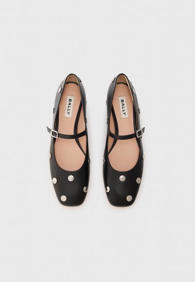 Black leather flat shoes with a square toe, adorned with large metallic studs, featuring a cross strap with a buckle detail.