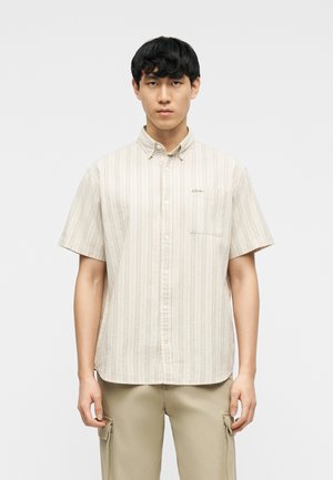 Junger Mann trägt ein beige gestreiftes Hemd mit kurzen Ärmeln und hellbraune Cargohosen, steht vor einem schlichten Hintergrund.