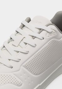 Baskets blanches avec panneau latéral perforé, lacets blancs passant à travers cinq œillets, et doublure intérieure grise visible.