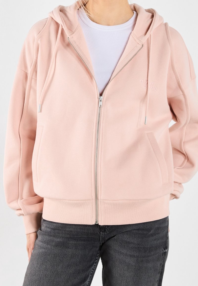 Femme portant un sweat à capuche zippé rose pâle avec cordons sur un t-shirt côtelé blanc et un jean gris foncé, debout avec une main dans la poche.