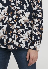 Marineblå skjorte med knapper, der har et hvidt og brunt abstrakt blomsterprint. Lavet af glat stof med lange ærmer og en afslappet pasform.