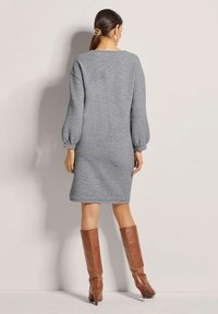 Graues Strickkleid mit langen Puffärmeln, knielang, runder Ausschnitt und lockerer Passform. Getragen mit braunen High Heels.