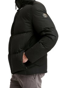 Veste matelassée noire avec un design matelassé, un col montant et un accent logo sur la manche gauche. Comprend des poches latérales zippées.