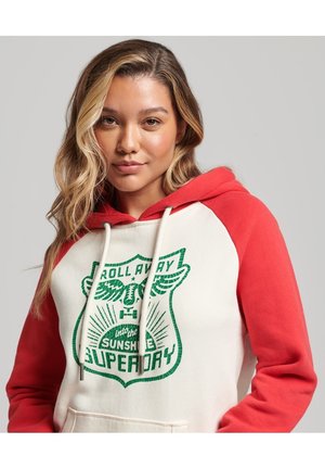 Jeune femme aux cheveux blonds ondulés portant un sweat à capuche blanc et rouge avec un texte graphique vert et une poche devant, sur un fond uni.