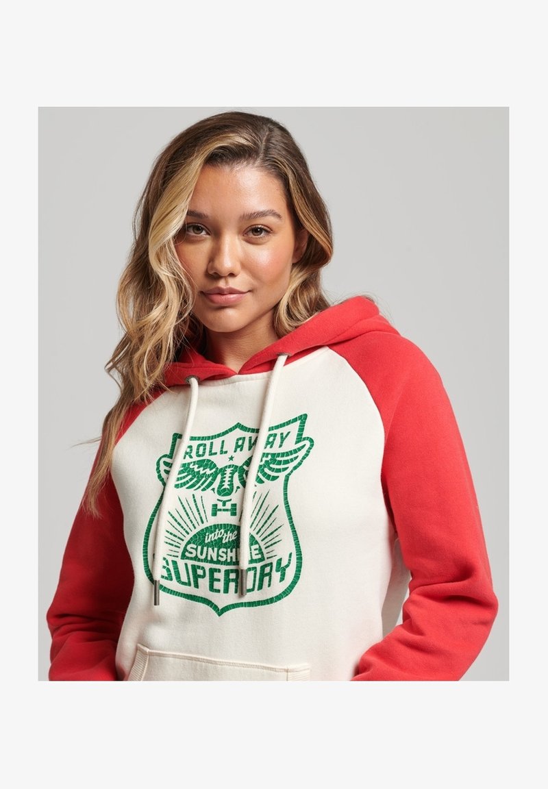 Jeune femme aux cheveux blonds ondulés portant un sweat à capuche blanc et rouge avec un texte graphique vert et une poche devant, sur un fond uni.