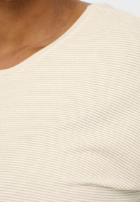 Gros plan sur une personne portant un haut crème en tissu côtelé avec de fines rayures horizontales et un col arrondi.